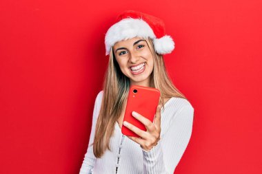 Güzel İspanyol kadın Noel şapkası takıyor. Elinde akıllı telefon var. Olumlu ve mutlu görünüyor. 