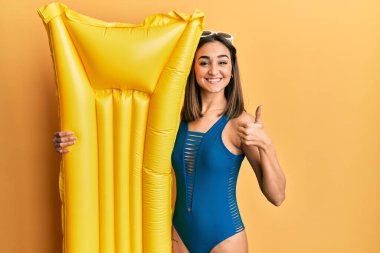 Genç esmer kız mayo giyiyor ve elinde şamandırayla gülümsüyor, mutlu ve pozitif, başparmağıyla mükemmel ve onay işareti yapıyor. 