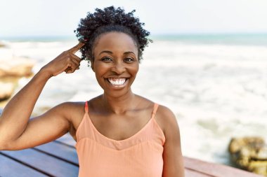 Genç Afrikalı Amerikalı kadın, yaz kıyafetleri içinde sahilde durup mutlu mesut parmakla işaret ediyor. 