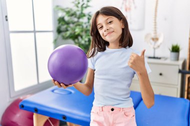 Genç İspanyol kız elinde küçük bir rehabilitasyon balosu tutuyor mutlu ve pozitif gülümsüyor, başparmağıyla mükemmel ve onay işareti yapıyor. 