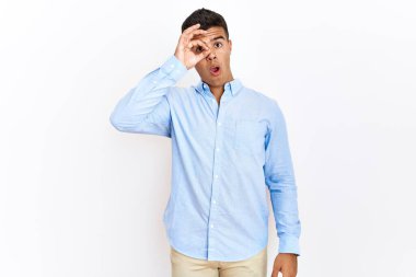 Soyutlanmış arka plan üzerinde duran İspanyol gömlekli genç adam şaşırmış yüz ifadesiyle şaşırmış, parmaklarının arasından bakan bir hareket yapıyor. İnançsız ifade. 