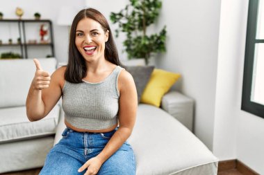 Evdeki kanepede oturan genç esmer kadın elleriyle mutlu bir jest yapıyor. Kameraya bakıp başarıyı gösteren ifade. 