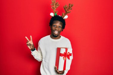 Young african american man wearing deer christmas hat holding gift smiling looking to the camera showing fingers doing victory sign. number two. 