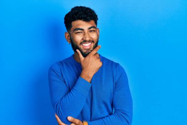 Sakallı, sıradan mavi süveter giyen, kameraya kendinden emin, kolları çapraz ve çenesi havada gülümseyen Arap bir adam. Olumlu düşünmek. 