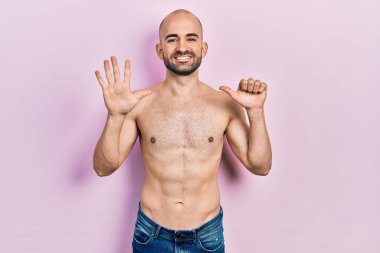 Genç kel adam üstsüz duruyor ve altı numaralı parmaklarıyla işaret ediyor kendinden emin ve mutlu bir şekilde gülümsüyor.. 