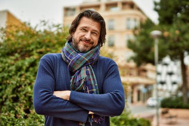 Orta yaşlı yakışıklı adam parkta kollarını kavuşturup gülümsüyor.