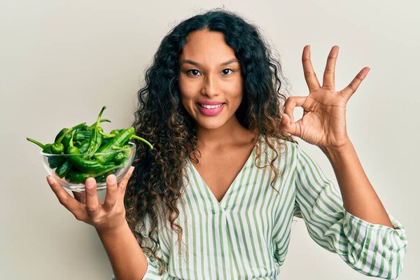 Young latin woman holding bowl with green peppers doing ok sign with fingers, smiling friendly gesturing excellent symbol 