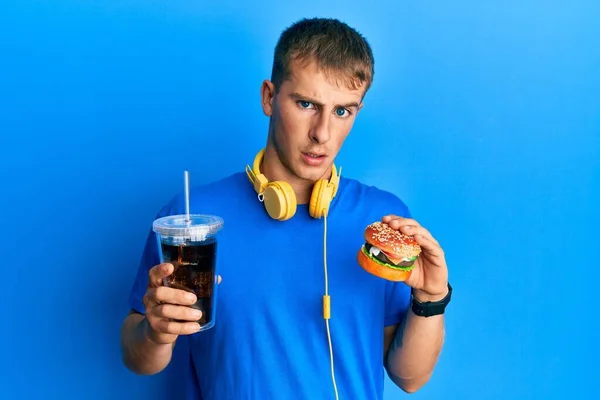 若い白人男性はおいしい古典的なバーガーとソーダ無知で混乱した表現を食べています 疑わしい概念 — ストック写真
