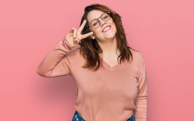 Genç ve cüsseli bir kadın günlük kıyafetler ve gözlük takıyor barış sembolü yapıyor. Yüzünde parmakları var, gülümsüyor ve zaferini gösteriyor. 