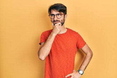 Gündelik kıyafetler giyen ve eli çenesinde gözlüklü soru soran genç İspanyol adam, düşünceli yüz ifadesi. Düşünceli bir yüzle gülümsüyor. şüphe kavramı. 