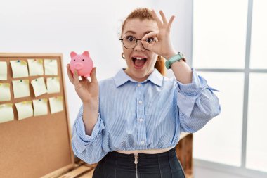 Genç kızıl saçlı kadın domuz kumbarasını elinde tutuyor gülümsüyor mutlu bir şekilde tamam işareti yapıyor parmaklarının arasından bakıyor 