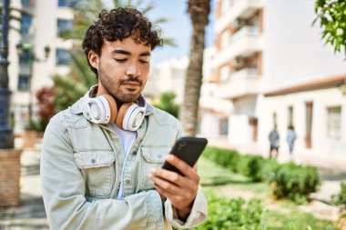 Yakışıklı, sakallı, güneşli bir günde mutlu bir şekilde gülümseyen kulaklık takan akıllı telefonlu müzik dinleyen bir İspanyol.