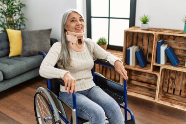 Orta yaşlı, gri saçlı, gülümseyen, kendine güvenen bir kadın, evinde kaza için tekerlekli sandalyede oturuyor.