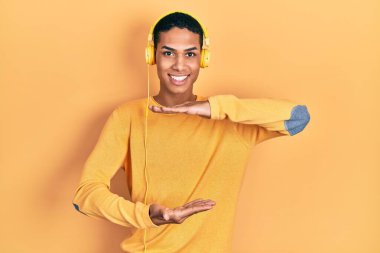 Genç Afrikalı Amerikalı adam müzik dinliyor kulaklıkla el kol hareketi yapıyor büyük ve büyük boy işareti, ölçü sembolü gösteriyor. Kameraya bakıp gülümsüyor. ölçüm kavramı. 