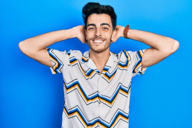 Genç İspanyol bir adam günlük kıyafetler giyiyor rahatlıyor ve esniyor, kolları ve elleri başının arkasında mutlu bir şekilde gülümsüyor. 