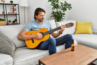 Orta yaşlı, beyaz, güler yüzlü, evde klasik gitar çalan bir adam.