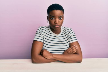 Genç Afro-Amerikan kadın, gündelik giysiler giyip şüpheli ve gergin bir şekilde masada oturuyor. Yüzünde çapraz kollarla onaylamayan bir ifade var. negatif kişi. 