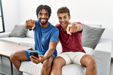 Genç ispanyol adamlar evdeki koltukta oturup seni ve kamerayı göstererek gülümsüyorlar. 