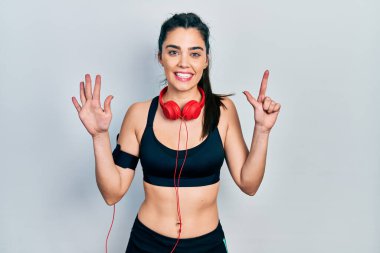 Spor kıyafetleri giyen, kulaklık takan genç İspanyol kız yedi numaralı parmaklarını gösterip işaret ederken kendinden emin ve mutlu bir şekilde gülümsüyor.. 
