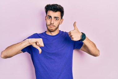 Gündelik tişört giyen genç İspanyol adam baş parmak kaldırıyor, anlaşmazlık ve anlaşma ifadesi takınıyor. çılgın çatışma 