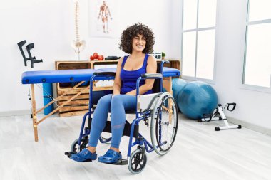 Fizyoterapi kliniğinde tekerlekli sandalyede oturan genç orta doğulu kadın kameraya seksi bir ifadeyle bakıyor, neşeli ve mutlu bir yüzle.. 