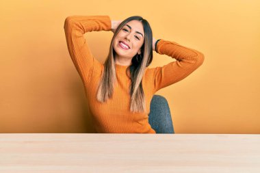 Latin kökenli genç bir kadın, gündelik giysiler içinde masada oturuyor, rahatlıyor ve esniyor, kolları ve elleri başının ve boynunun arkasında mutlu bir şekilde gülümsüyor. 