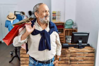 Butik dükkanında alışveriş torbaları tutan yakışıklı bir adam yüzünde gülümseme ve doğal bir ifadeyle yan yana bakıyor. Kendinden emin gülen. 
