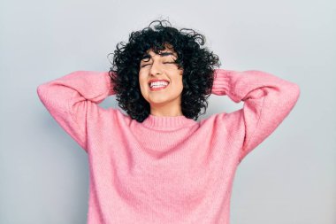 Orta Doğulu genç bir kadın günlük kıyafetler giyiyor, rahatlıyor ve esniyor, kolları ve elleri başının arkasında mutlu bir şekilde gülümsüyor. 