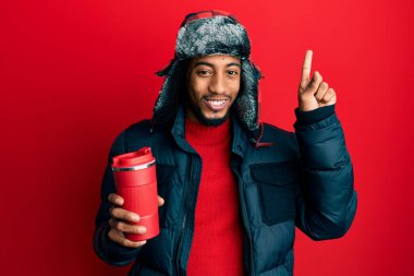 Sakallı, kış şapkalı, termosu üzerinde sıcak kahve tutan ve gülümseyen genç Afrikalı Amerikalı bir adam bir fikir ya da soruyla gülümseyen mutlu yüzlü, bir numara. 