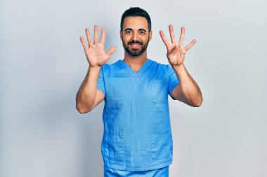 Sakallı yakışıklı, mavi erkek hemşire üniforması giyen, dokuz numaralı parmaklarıyla işaret eden, kendinden emin ve mutlu bir adam.. 