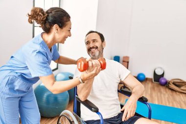 Orta yaşlı bir adam ve kadın, fizyoterapi kliniğinde tekerlekli sandalyede oturan dambılı kullanarak rehabilitasyon seansı yapıyorlar.