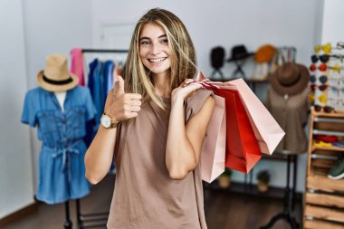 Perakende dükkanında alışveriş torbaları tutan genç sarışın kadın elleriyle mutlu bir jest yapıyor. Kameraya bakıp başarıyı gösteren ifade. 