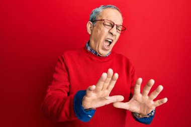 Gri saçlı, günlük kıyafetler ve gözlük takan yakışıklı, tiksindirici ve tiksindirici bir yüz ifadesi takınan, tiksinti reaksiyonu yüzünden tiksinmiş ve korkmuş bir adam. Eller havada. 