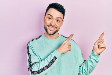 Gündelik kıyafetler giyen genç İspanyol adam gülümseyip kameraya bakıyor ve iki eliyle ve parmaklarıyla kenarı işaret ediyor.. 