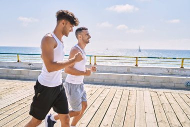 İki İspanyol erkek, sportif, gülümseyen bir çift, deniz kenarında koşuyorlar.