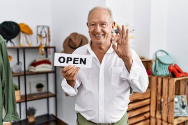 Perakende dükkanında elinde açık metinle afiş tutan kıdemli bir adam tamam işareti yapıyor parmaklarıyla gülümseyerek mükemmel bir sembol oluşturuyor. 