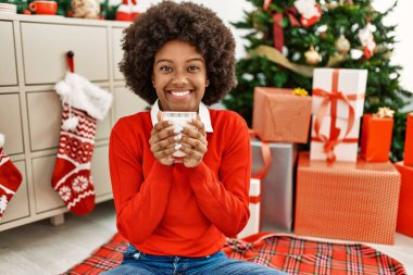Genç Afrikalı Amerikalı kadın evde Noel ağacının yanında süt içiyor.
