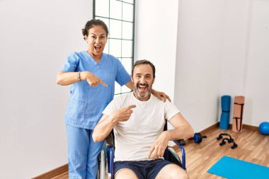 İspanyol orta yaşlı bir adam tekerlekli sandalyede oturuyor ve rehabilitasyon kliniğinde hemşire mutlu bir şekilde elini ve parmağını göstererek gülümsüyor. 