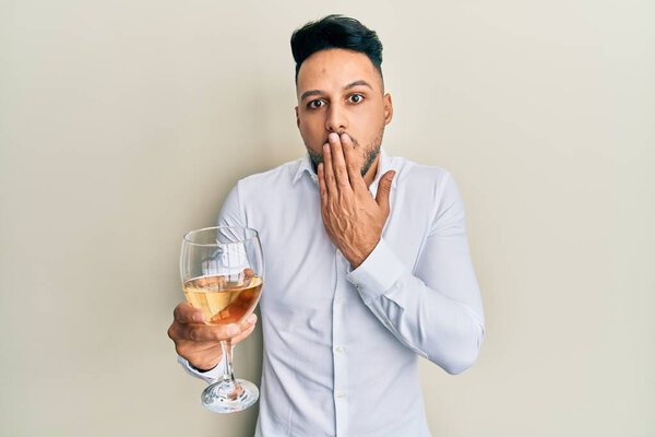 Young arab man drinking a glass of white wine covering mouth with hand, shocked and afraid for mistake. surprised expression 