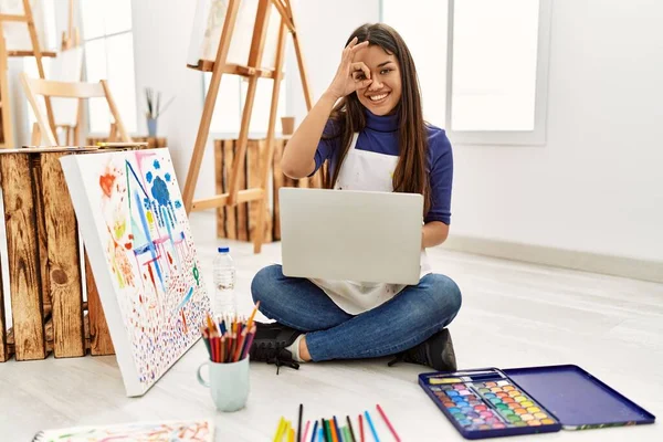 年轻的黑发女人坐在艺术工作室的地板上 带着笔记本电脑 开心地微笑着 手拿着手签 用手指看着 — 图库照片