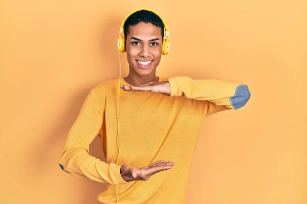 年轻的非洲裔美国人一边听音乐 一边用耳机做手势 手举着大号和大号的标志 测量符号 微笑着看着摄像机 计量概念 — 图库照片