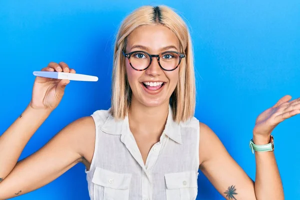 Beautiful blonde woman holding pregnancy test result celebrating achievement with happy smile and winner expression with raised hand