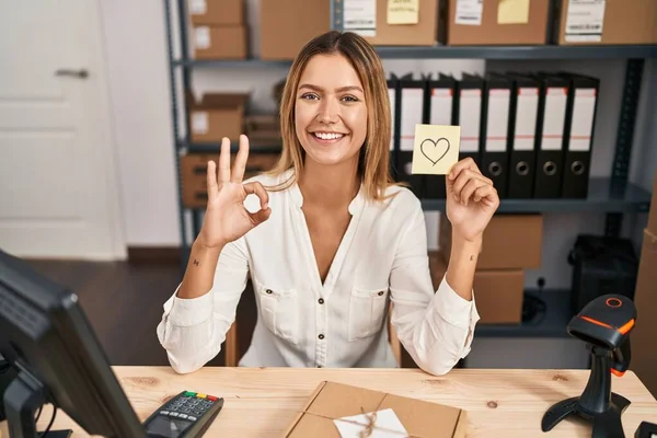 Young blonde woman working at small business ecommerce holding heart note doing ok sign with fingers, smiling friendly gesturing excellent symbol