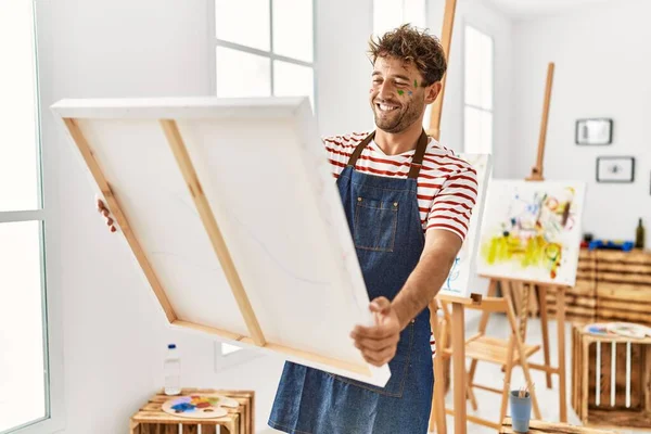 年轻的惊慌失措的男人带着自信的笑容在艺术工作室里举着画布 — 图库照片
