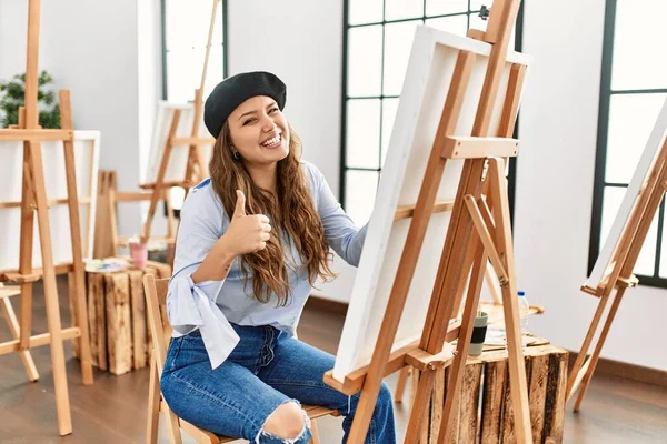 若いヒスパニックアーティストの女性は アートスタジオでキャンバスに絵を描く手で積極的なジェスチャーを行うことを承認し 親指を笑顔と成功のために幸せ 勝者のジェスチャー — ストック写真