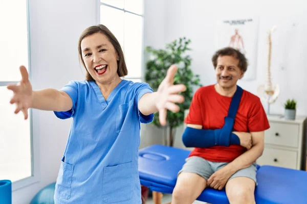 Middle age doctor woman with patient with arm injury at rehabilitation clinic looking at the camera smiling with open arms for hug. cheerful expression embracing happiness.