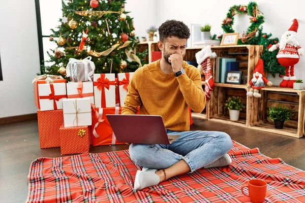 阿拉伯年轻人坐在圣诞树旁 用笔记本电脑闻到一股难闻难闻的难闻气味 用手指捂住鼻子呼吸 — 图库照片