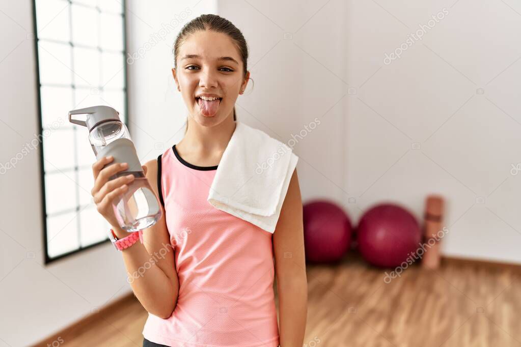 Young brunette teenager wearing sportswear holding water bottle sticking tongue out happy with funny expression. emotion concept. 
