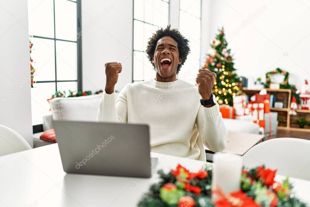 Young african american man using laptop sitting on the table by christmas tree very happy and excited doing winner gesture with arms raised, smiling and screaming for success. celebration concept. 