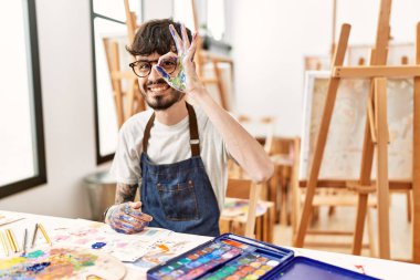 Resim stüdyosunda sakallı İspanyol bir adam el hareketleriyle gülümsüyor ve mutlu yüzüyle parmaklarının arasından bakıyor.. 